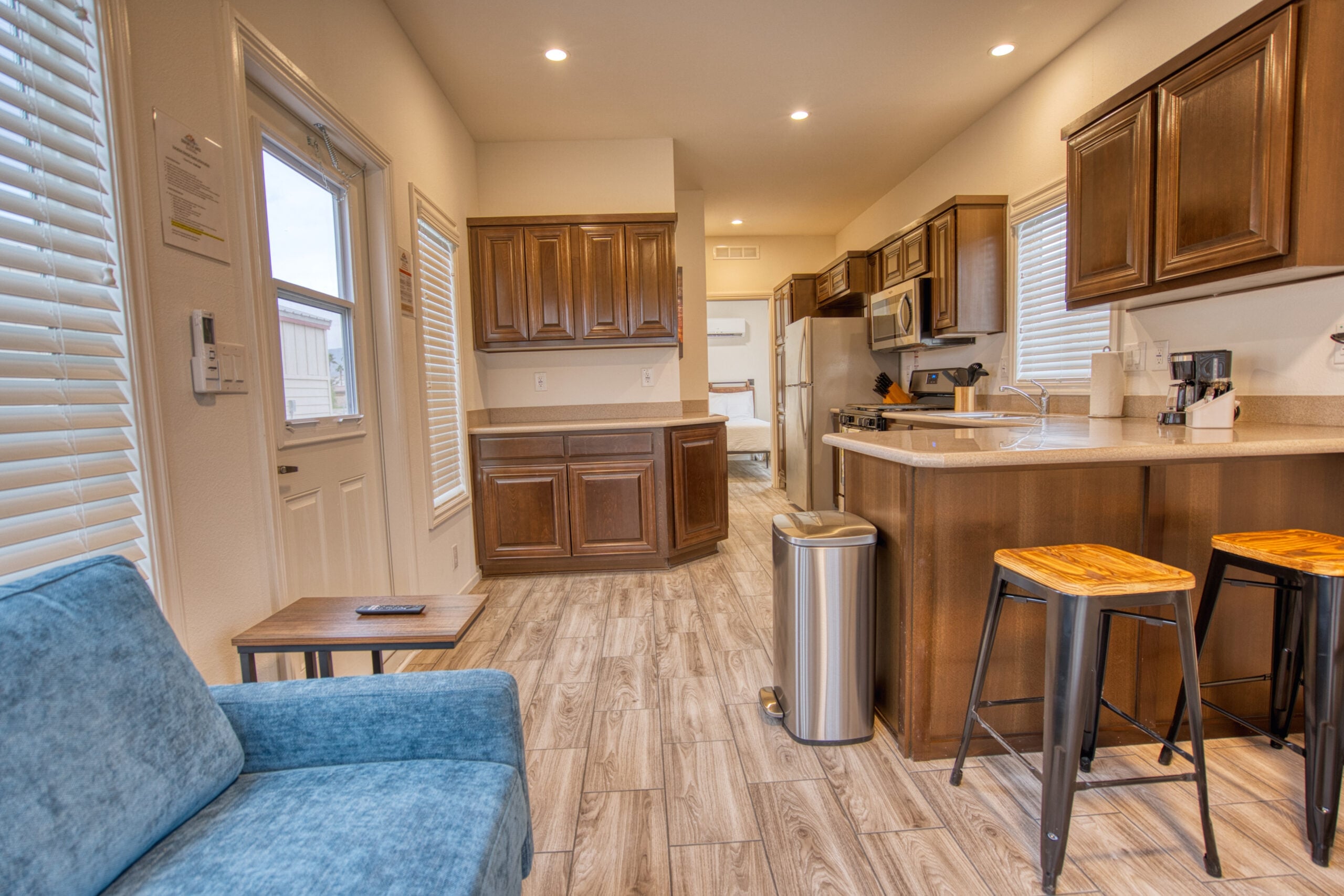 Modern kitchen and living area with wooden cabinets, stainless steel appliances, bar stools, blue sofa, and wood-look tile flooring.