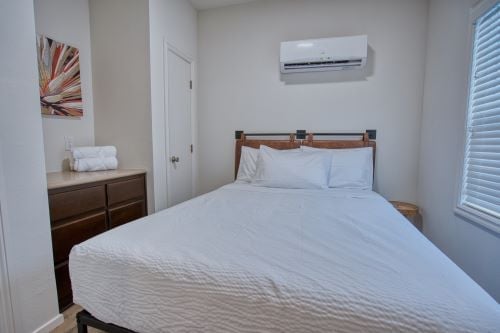 A neatly made bed with white linens is positioned against a wall under an air conditioner; a dresser with towels and artwork are to the left, and a window with blinds is on the right.