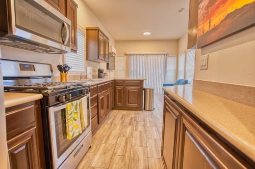 Modern kitchen with stainless steel appliances, brown cabinets, tan countertops, and wood-look tile flooring; sunlight enters through sliding glass doors in the background.