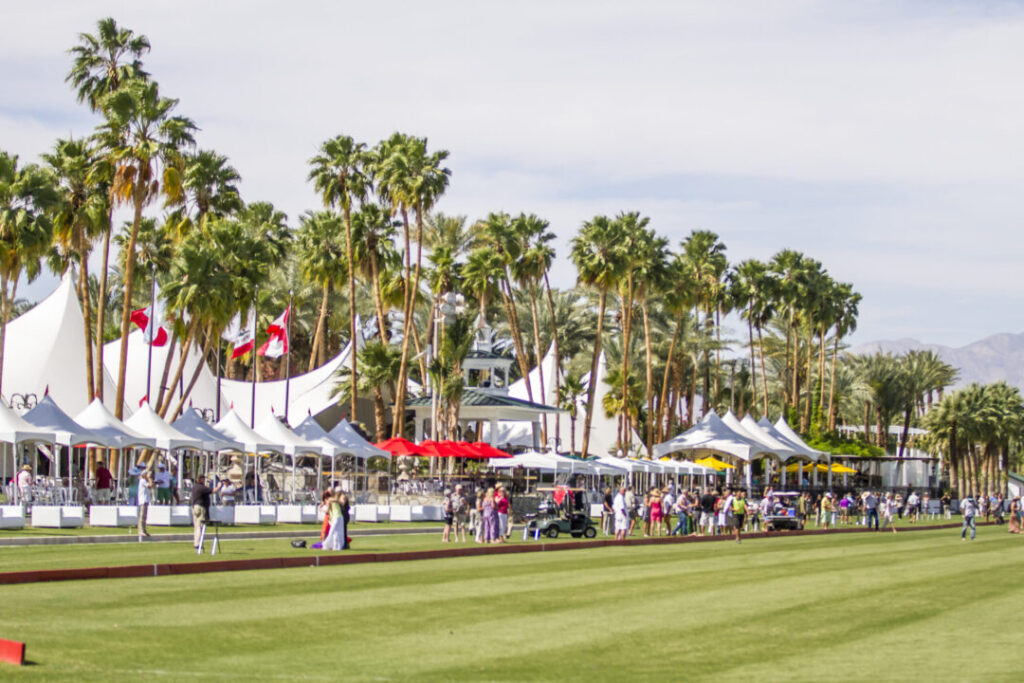 People gather along the edge of a polo field lined with palm trees and white tents under a partly cloudy sky.