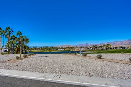 A gravel lot with sparse plants overlooks a lake, green grass, and palm trees, with mountains and a clear blue sky in the background.