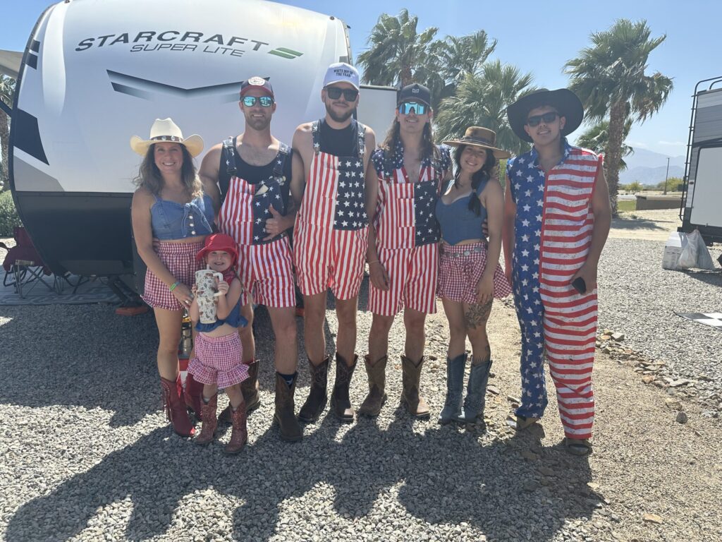 Seven people, including a child, pose outdoors in front of a camper, wearing red, white, and blue American flag-themed outfits and sunglasses.