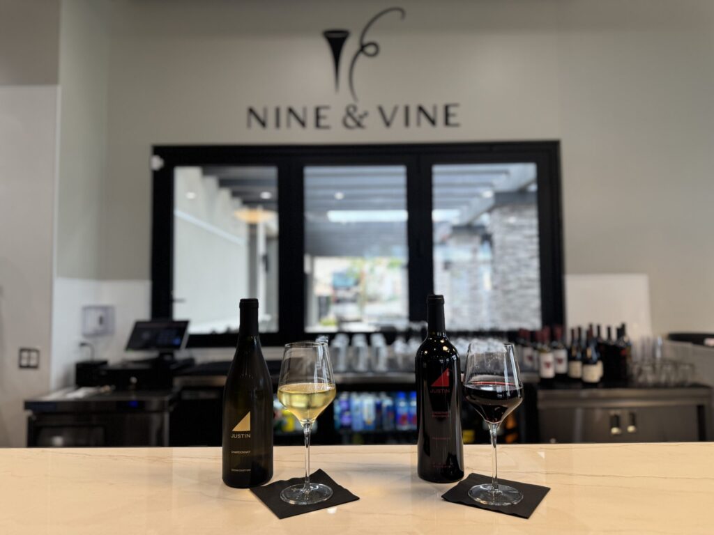 Two wine bottles and filled glasses, one white and one red, are displayed on a bar counter at Nine & Vine, with shelves of wine bottles visible in the background.