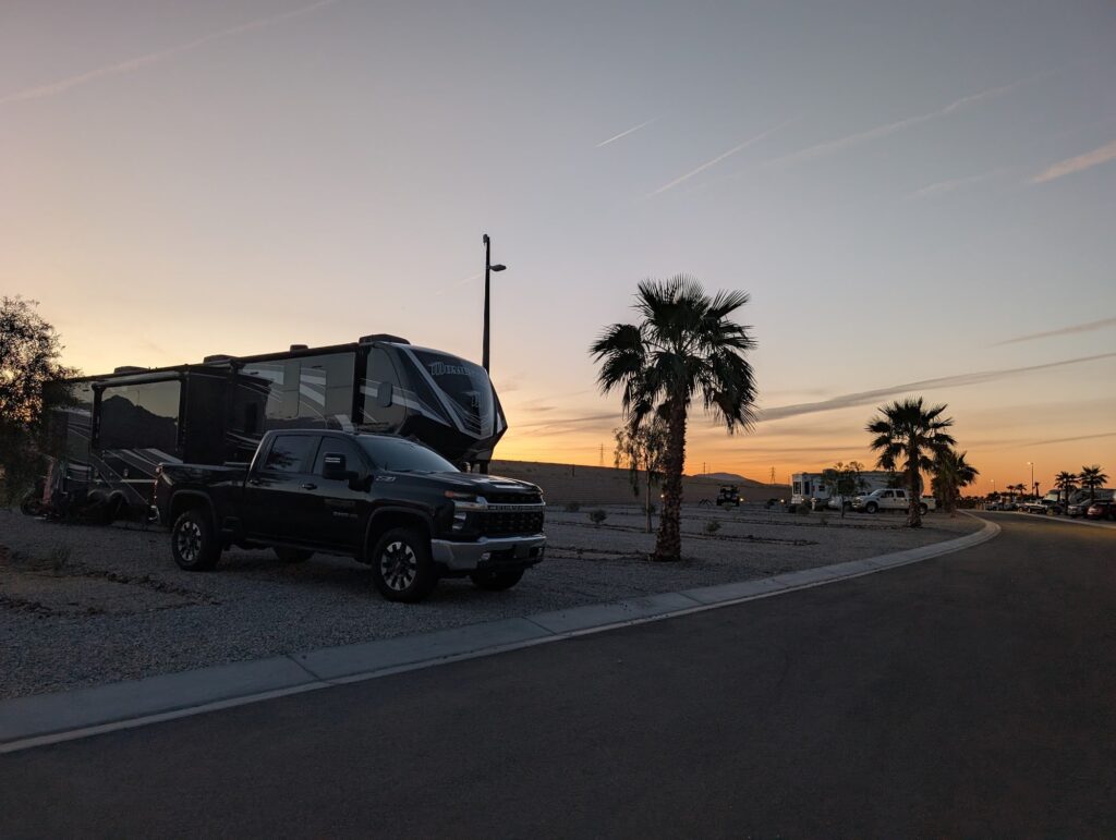 A black pickup truck and a large RV are parked on gravel near palm trees at a campground during sunset.