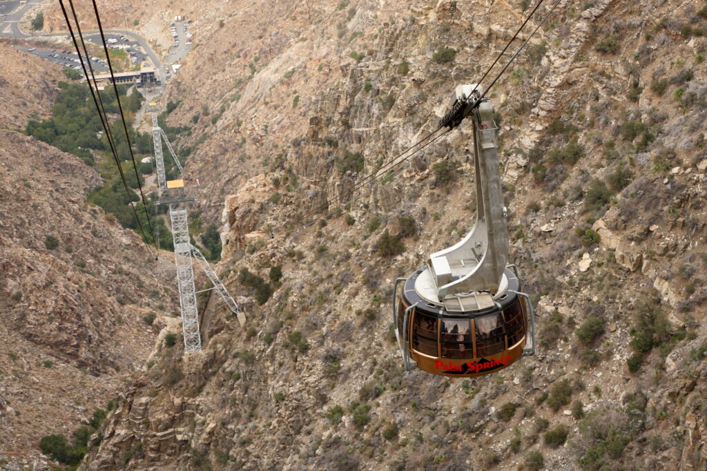 A round tram car labeled "Palm Springs" travels on cables over a rocky, mountainous landscape with towers and a valley below.