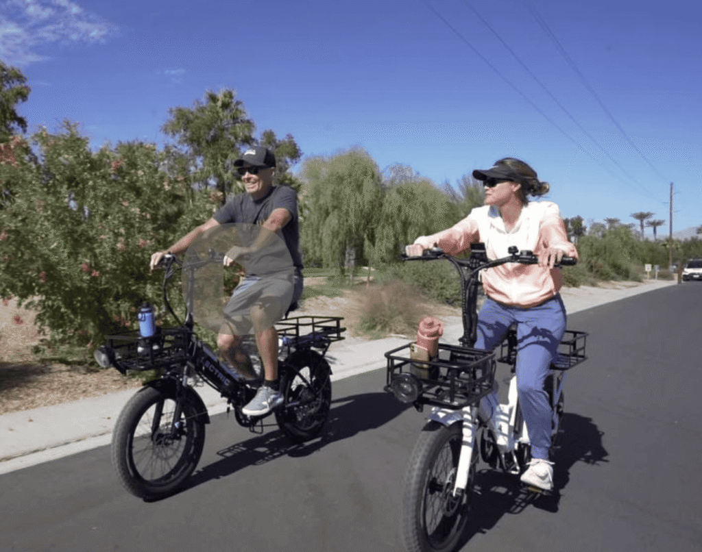 Two people ride electric bikes on a sunny street, both wearing sunglasses and casual clothing, with trees and blue sky in the background.