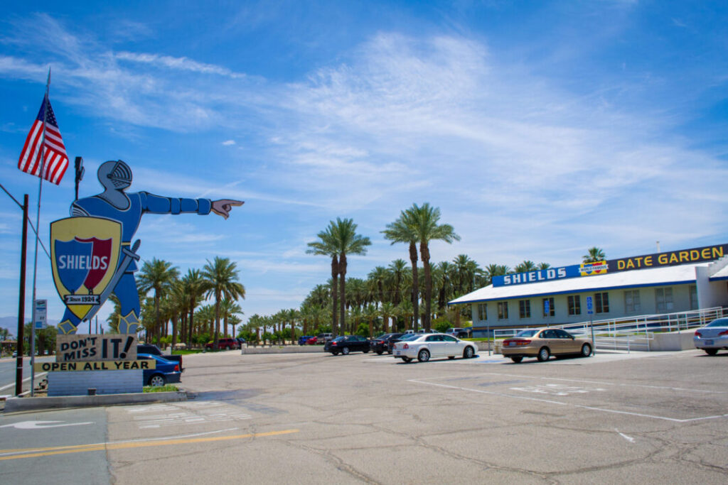 Parking lot with a large knight-shaped "Shields Date Garden" sign, palm trees, cars, and the Shields Date Garden building under a blue sky.
