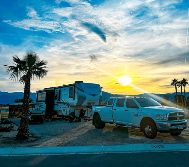 A white pickup truck and camper trailer are parked on a gravel lot with a palm tree nearby, mountains in the background, and the sun setting in a partly cloudy sky.