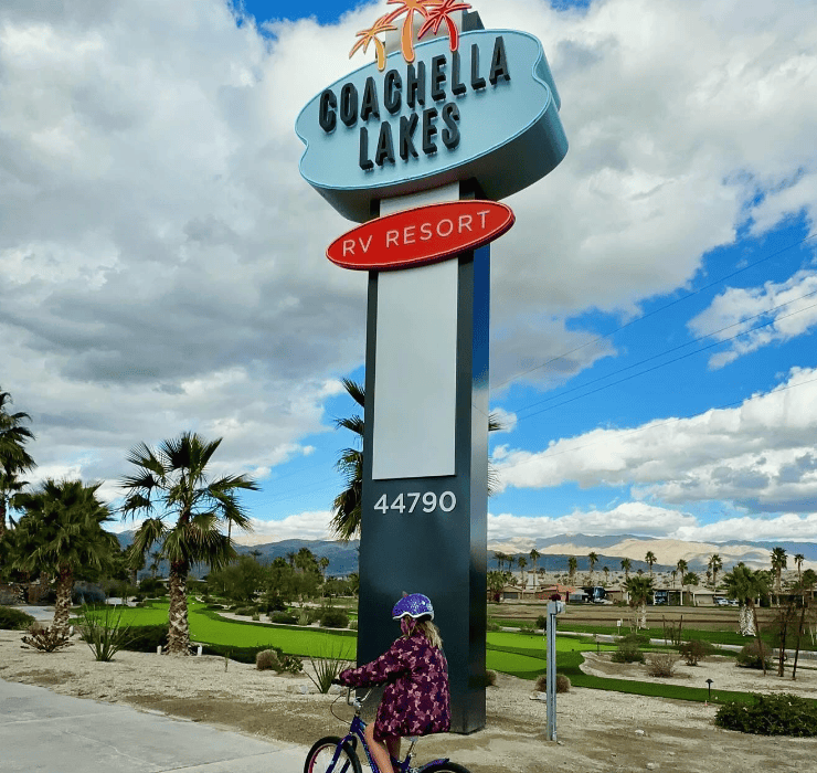 A child rides a bicycle near the Coachella Lakes RV Resort sign with palm trees and mountains in the background.