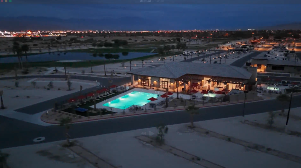 Aerial view of a modern building with an illuminated outdoor swimming pool at dusk, surrounded by desert landscape and distant city lights.
