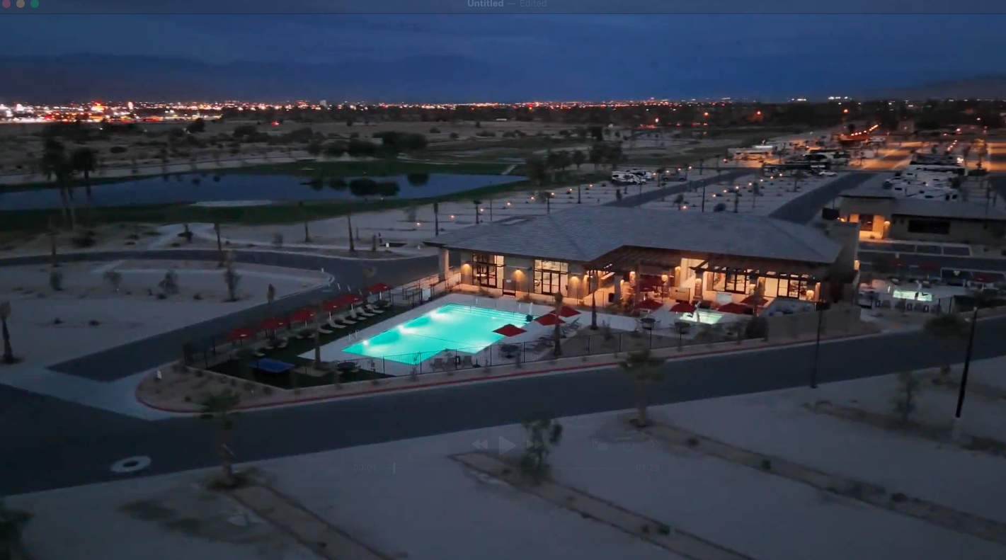 Aerial view of a modern building with an illuminated outdoor swimming pool at dusk, surrounded by desert landscape and distant city lights.