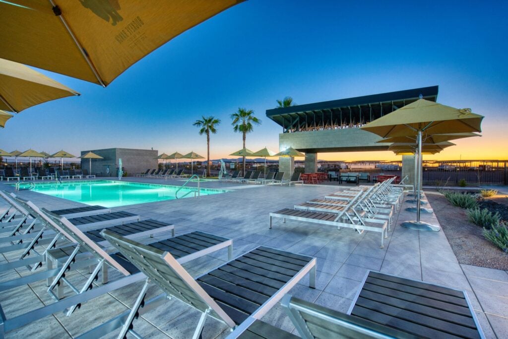 Outdoor swimming pool area at sunset with lounge chairs, yellow umbrellas, palm trees, and a modern building in the background.
