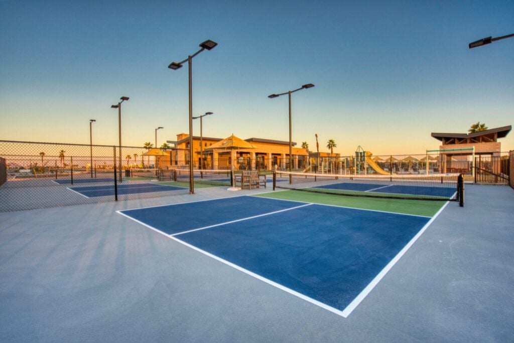 Outdoor pickleball courts with blue and green surfaces, surrounded by fencing, with a clubhouse and playground visible in the background at sunset.