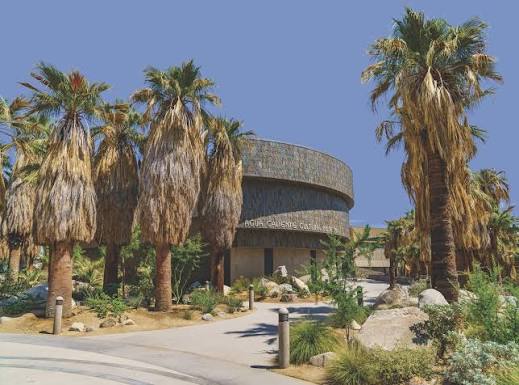 A modern building with a circular facade is surrounded by tall palm trees and desert landscaping under a clear blue sky.