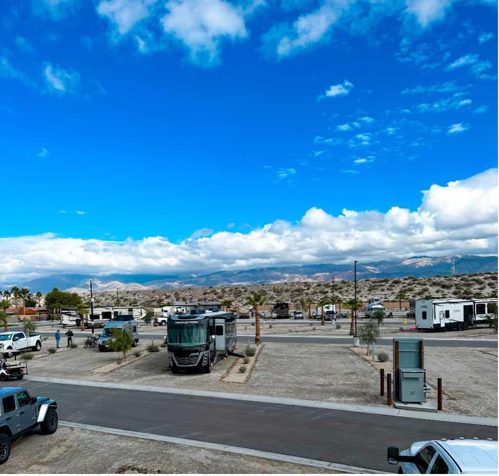 A paved RV park with several parked motorhomes and vehicles under a blue sky with scattered clouds, surrounded by desert landscape and distant mountains.