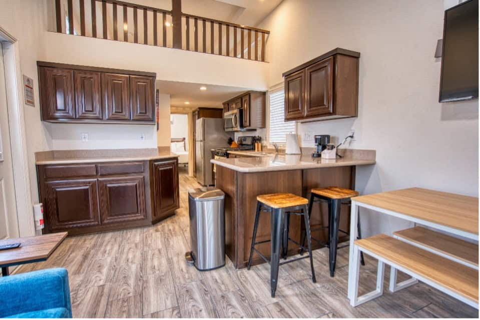 Modern kitchen with dark wood cabinets, an island with two stools, a dining table with benches, and a loft area above. Light wood flooring throughout.