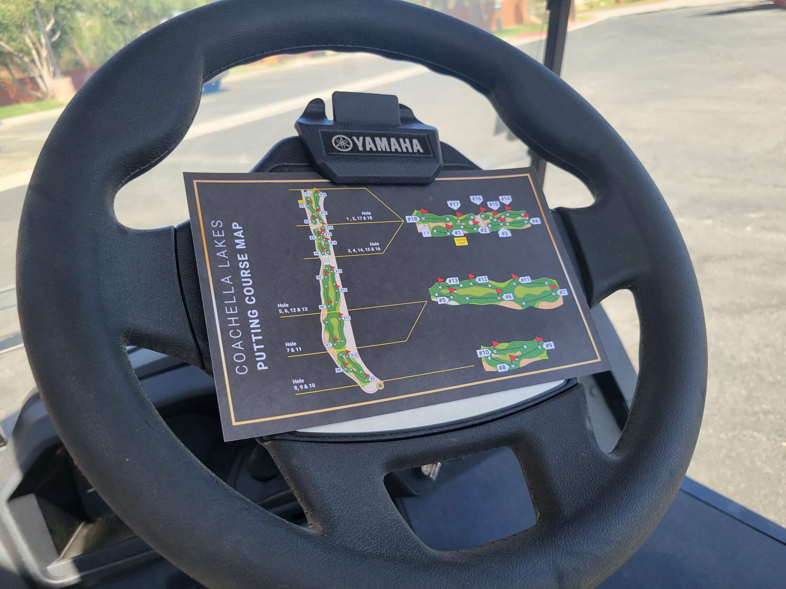 A golf cart steering wheel holds a clipboard with a Coachella Lakes putting course map, showing hole layouts and course details.
