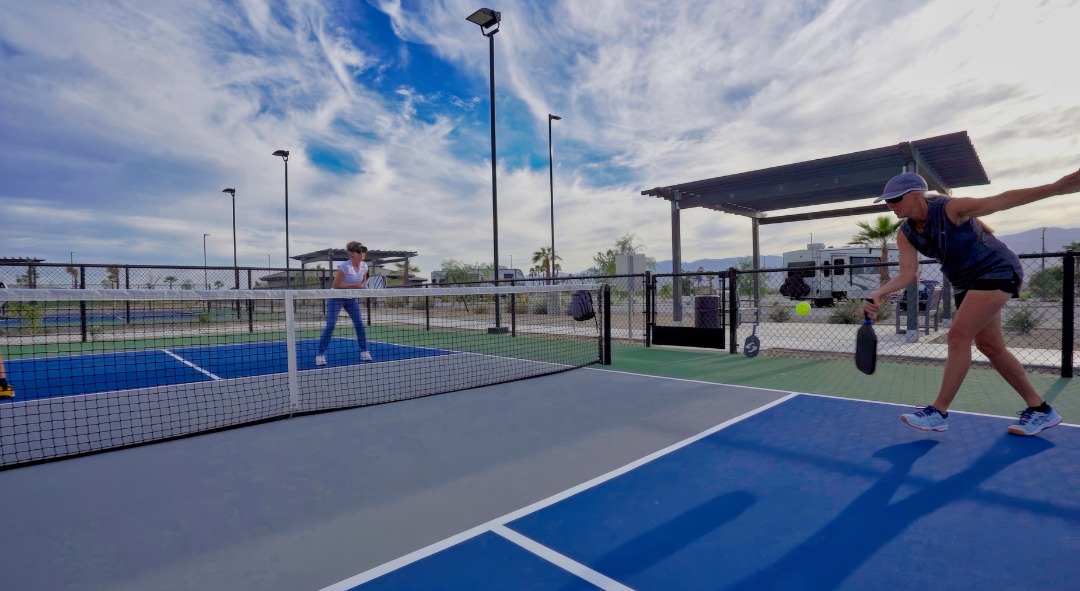 Two people play pickleball on an outdoor court under a partly cloudy sky, with one player preparing to hit the ball.