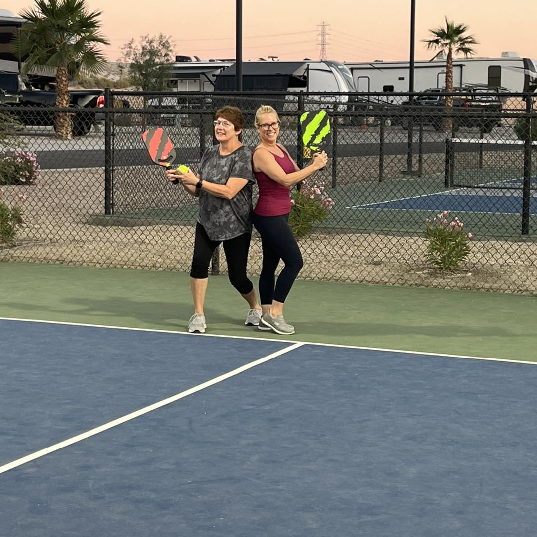 Two women stand back to back on an outdoor pickleball court, each holding a paddle, smiling at the camera. RVs and palm trees are visible in the background.
