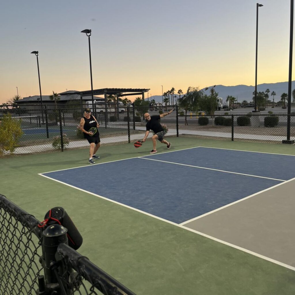 Two people play pickleball on an outdoor court at sunset, with palm trees, mountains, and buildings visible in the background.