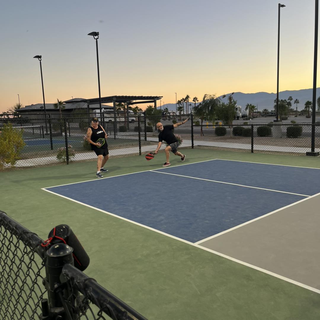 Two people play pickleball on an outdoor court at sunset, with palm trees, mountains, and buildings visible in the background.