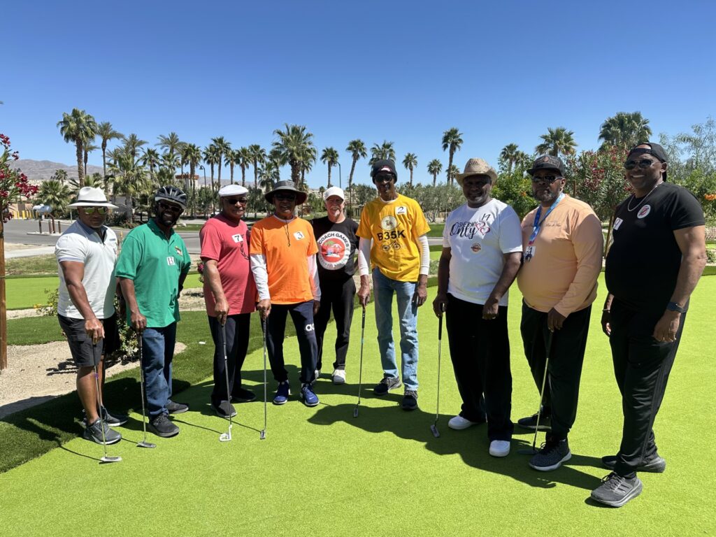 Nine men stand on a putting green holding golf clubs, with palm trees and clear blue sky in the background.