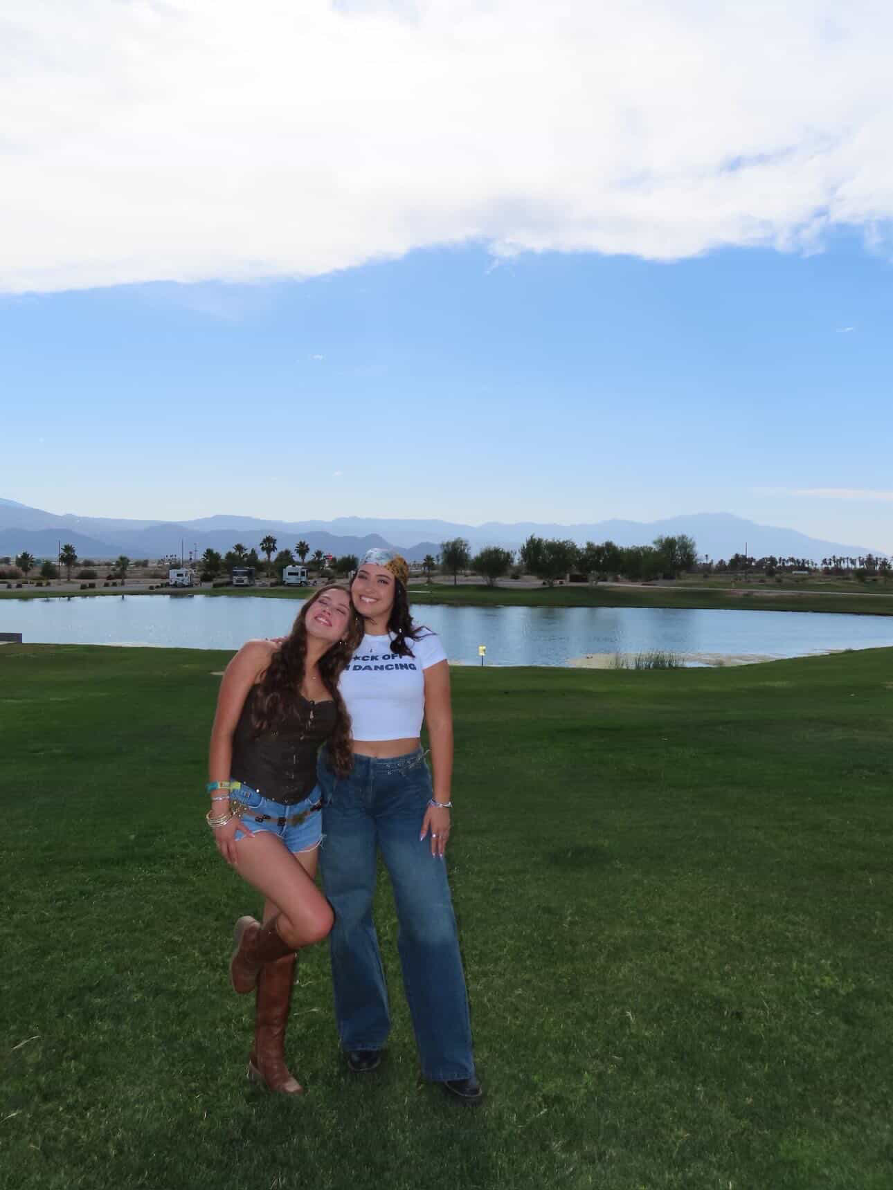 Two women pose and smile on a grassy field by a pond, with mountains and trees in the background under a partly cloudy sky.