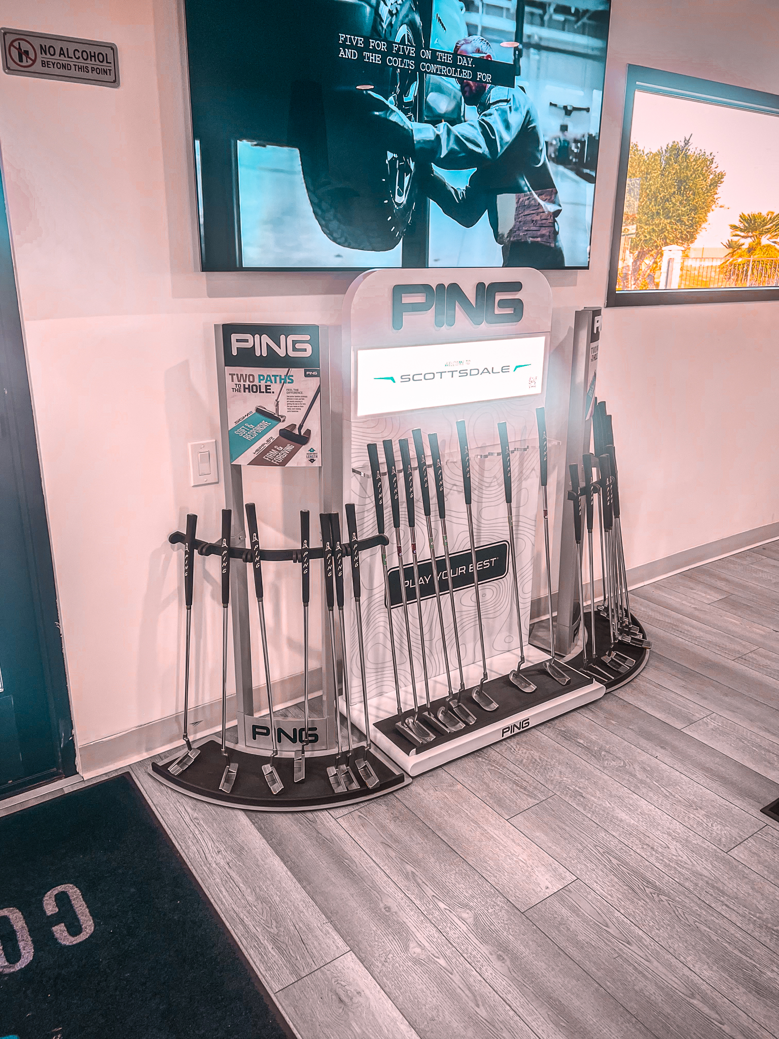 A display rack holds multiple Ping golf putters in a store, with signage above and a monitor showing golf equipment in the background.