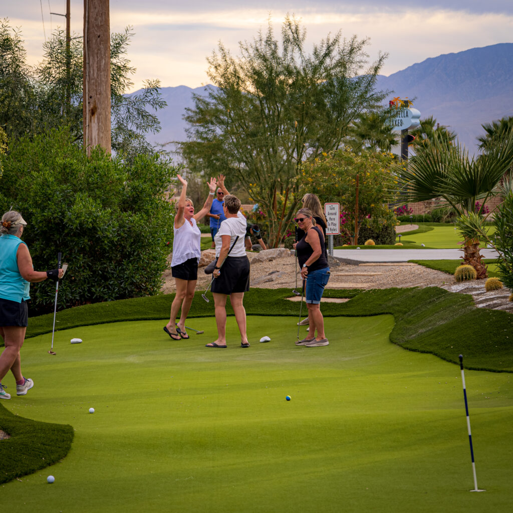 Five people are on a putting green; two are high-fiving, while others stand nearby with golf clubs. Desert scenery and mountains are visible in the background.