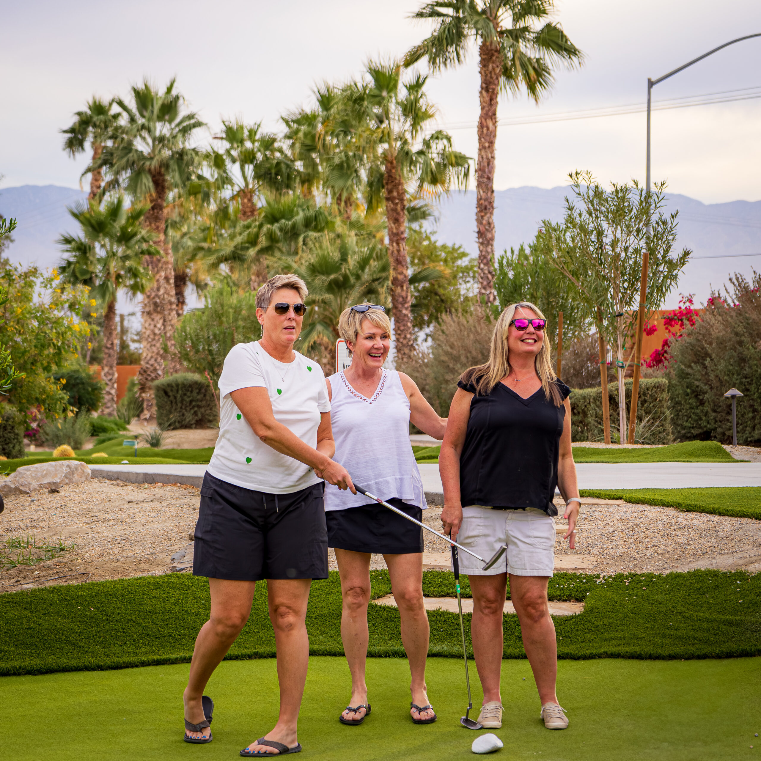 Three women stand on a golf course, smiling and holding golf clubs, with palm trees and mountains in the background.