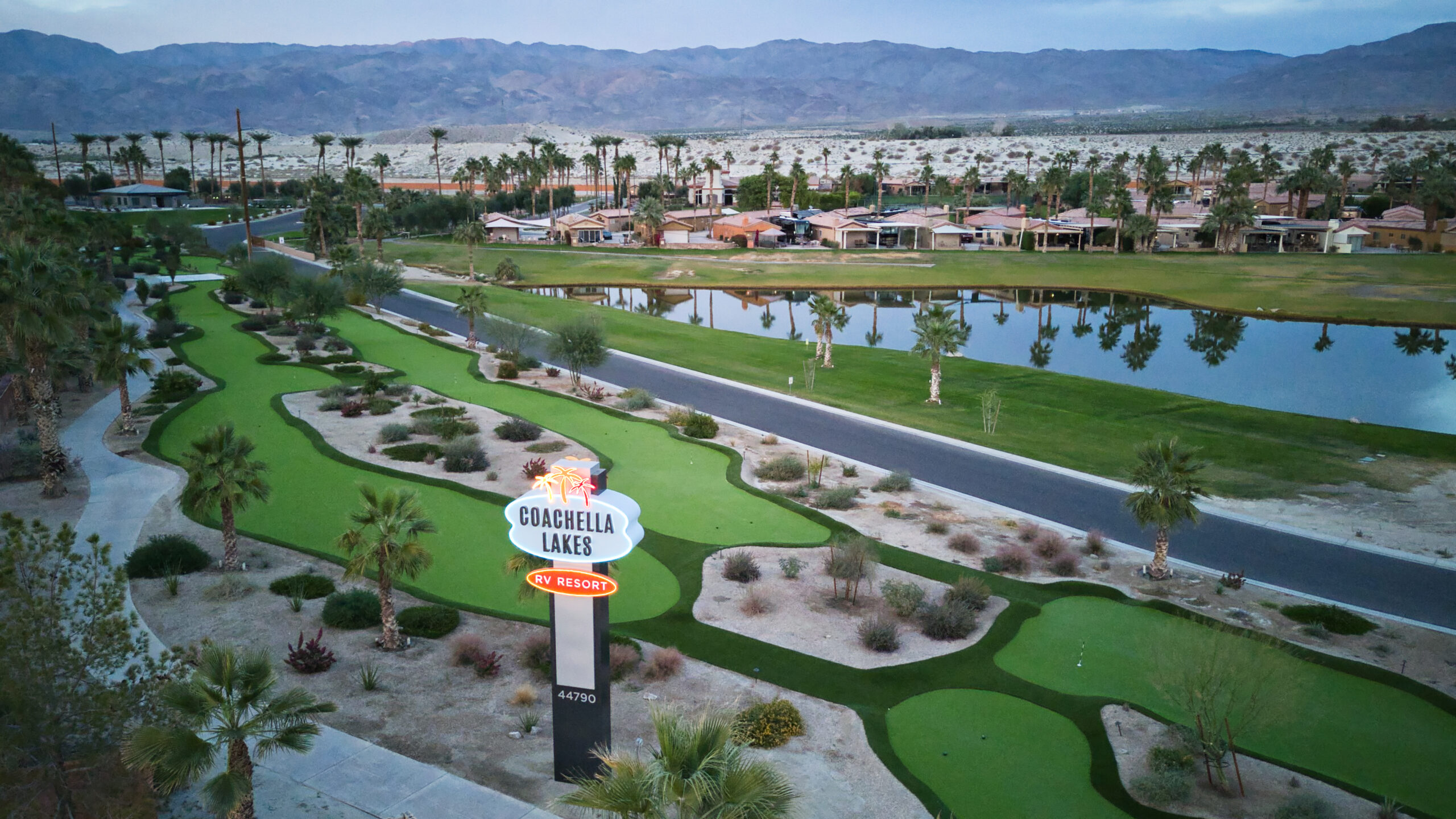 Aerial view of Coachella Lakes RV Resort sign with manicured landscaping, palm trees, pond, road, and houses set against desert mountains.
