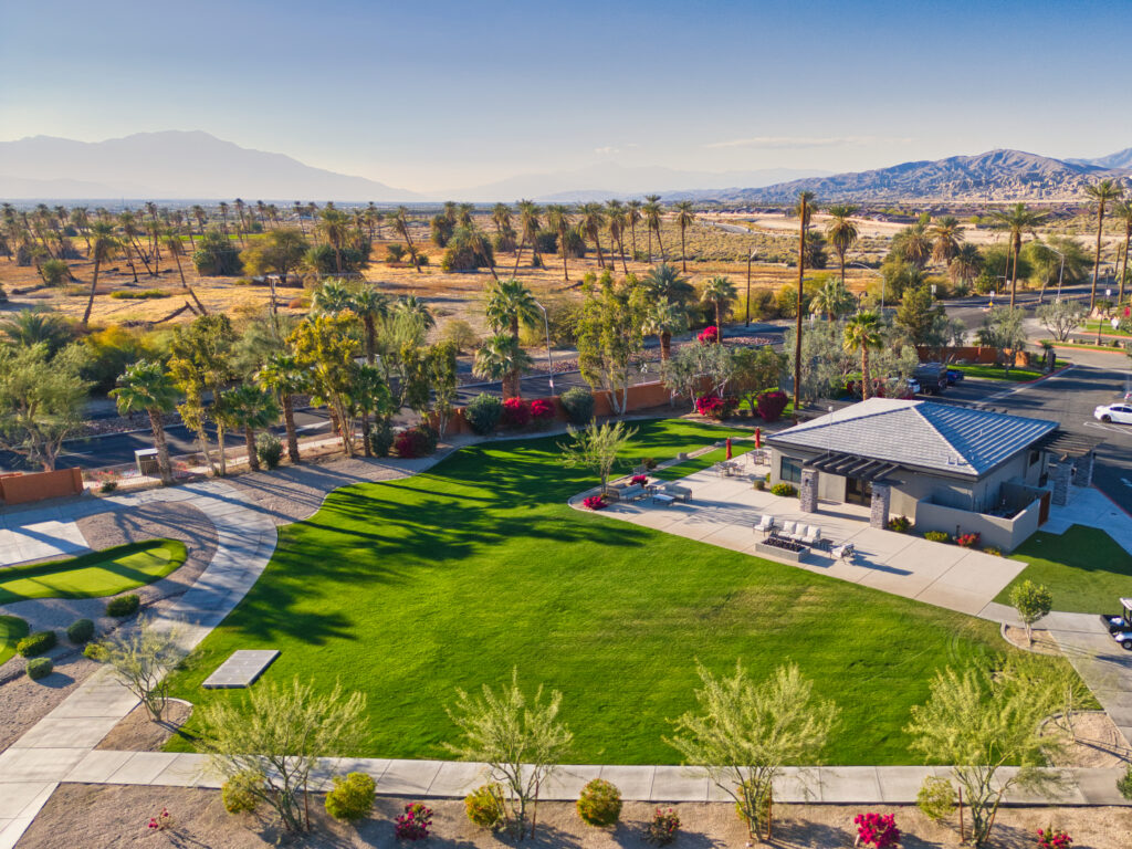 A modern building sits beside a green lawn and paths, surrounded by palm trees and desert landscape, with mountains visible in the background under a clear sky.