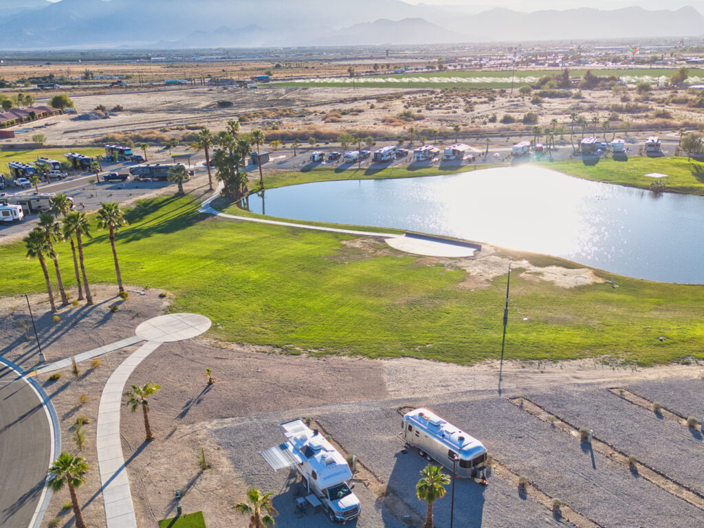 Aerial view of an RV park with a small pond, green grass, palm trees, and rows of parked RVs under clear skies in a desert landscape.