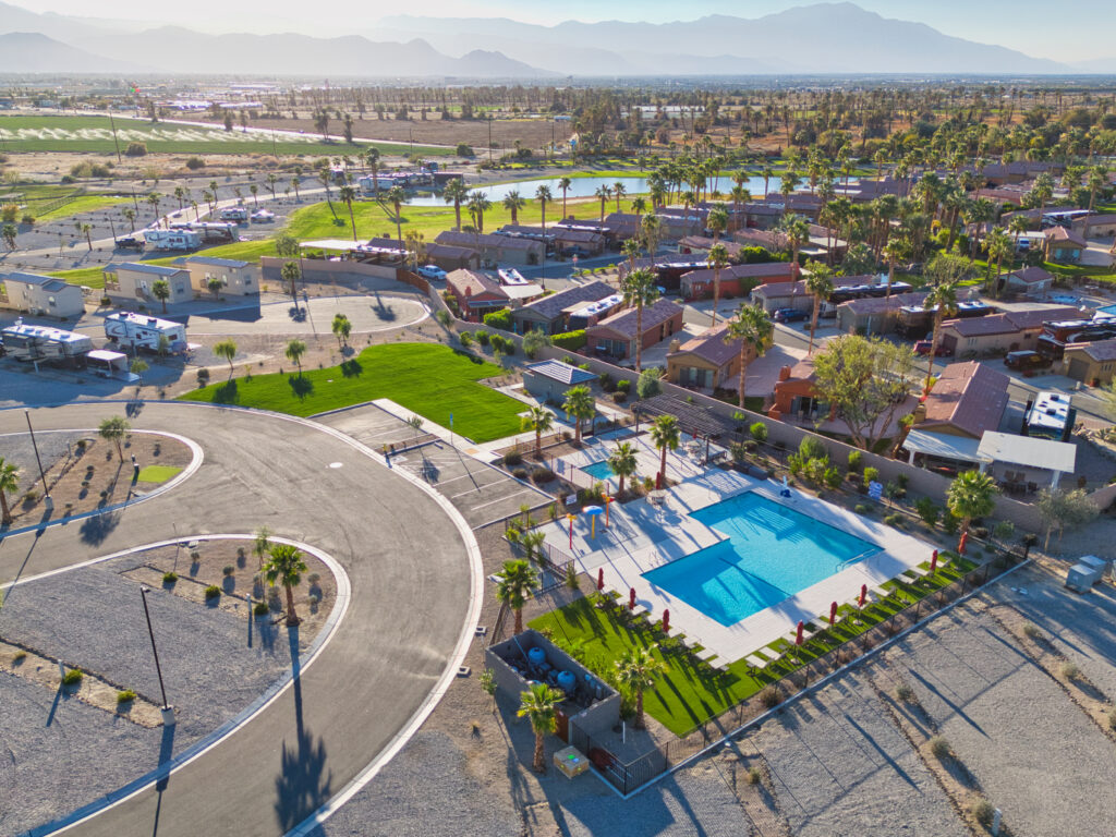 Aerial view of a residential neighborhood with a large swimming pool, palm trees, RVs, and mountains in the background under clear skies.