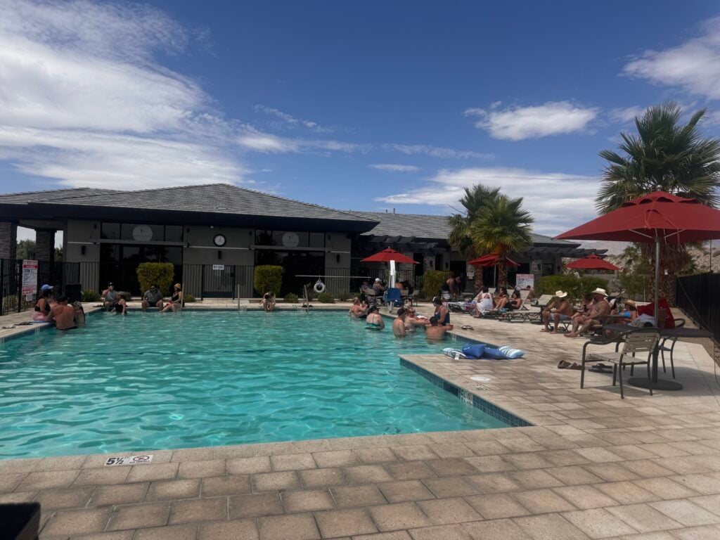 People relax and swim in an outdoor pool surrounded by lounge chairs, umbrellas, palm trees, and a building under a partly cloudy sky.