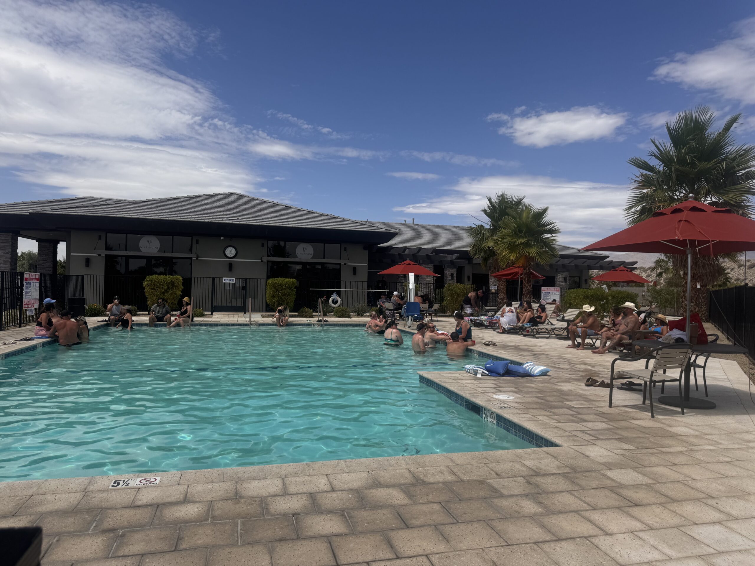 People relax and swim in an outdoor pool surrounded by lounge chairs, umbrellas, palm trees, and a building under a partly cloudy sky.