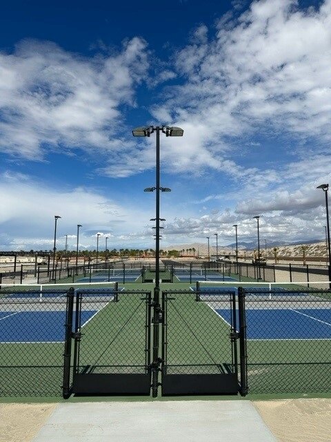 A fenced entrance leads to multiple empty tennis courts under a partly cloudy sky, with light poles lining the area.