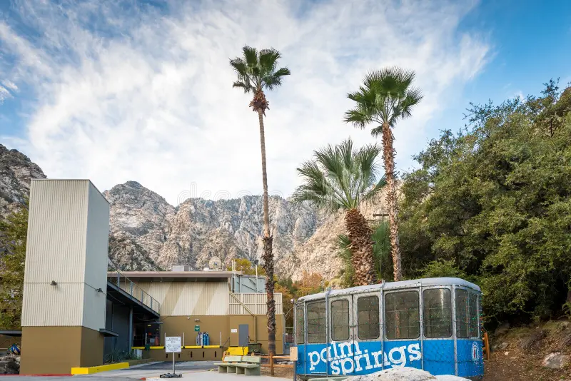 An old Palm Springs tram car is on display near palm trees, with a modern building and rocky mountains in the background under a partly cloudy sky.
