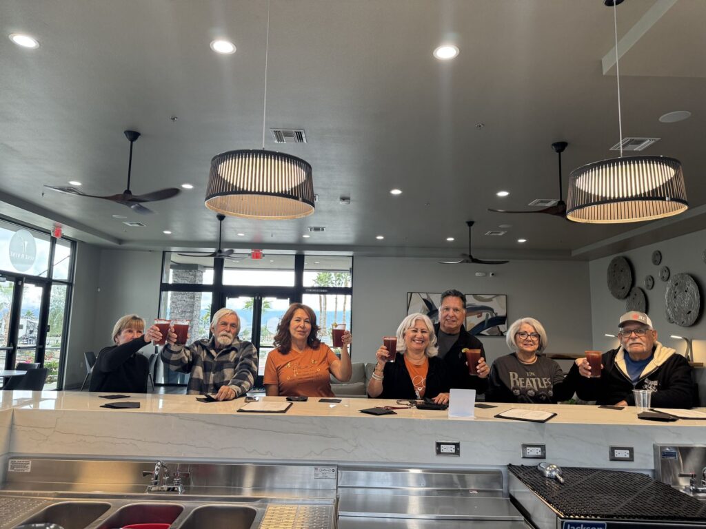 Seven people sitting at a countertop inside a modern cafe, raising glasses and smiling at the camera, with hanging lights and wall clocks in the background.