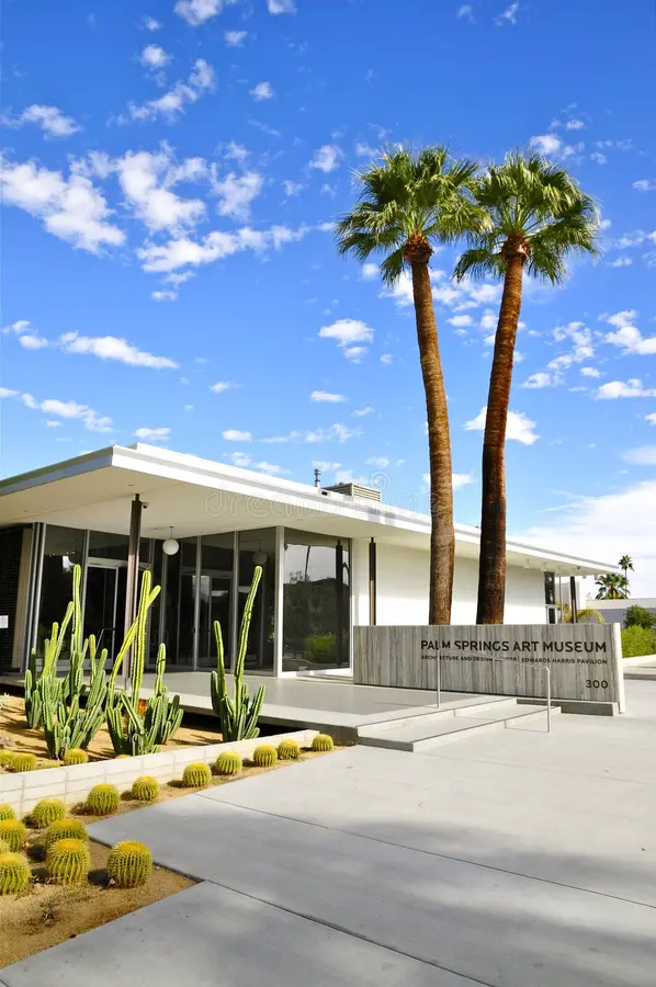 Modern building of the Palm Springs Art Museum surrounded by palm trees and desert landscaping under a blue sky with scattered clouds.