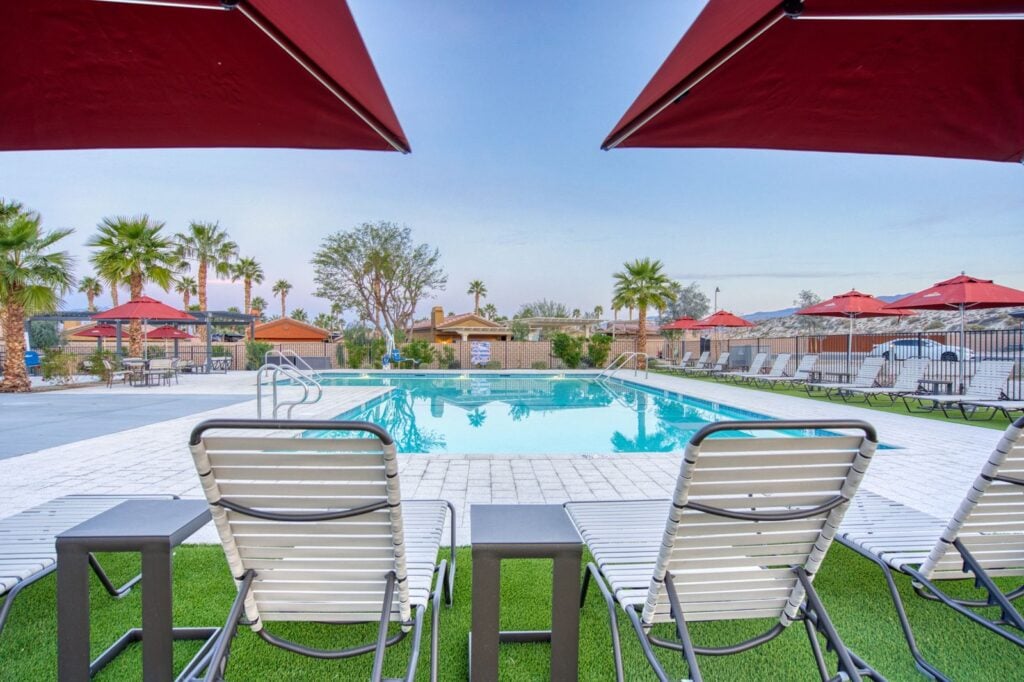 Three lounge chairs and small tables face a clear outdoor swimming pool with palm trees, red umbrellas, and blue sky in the background.