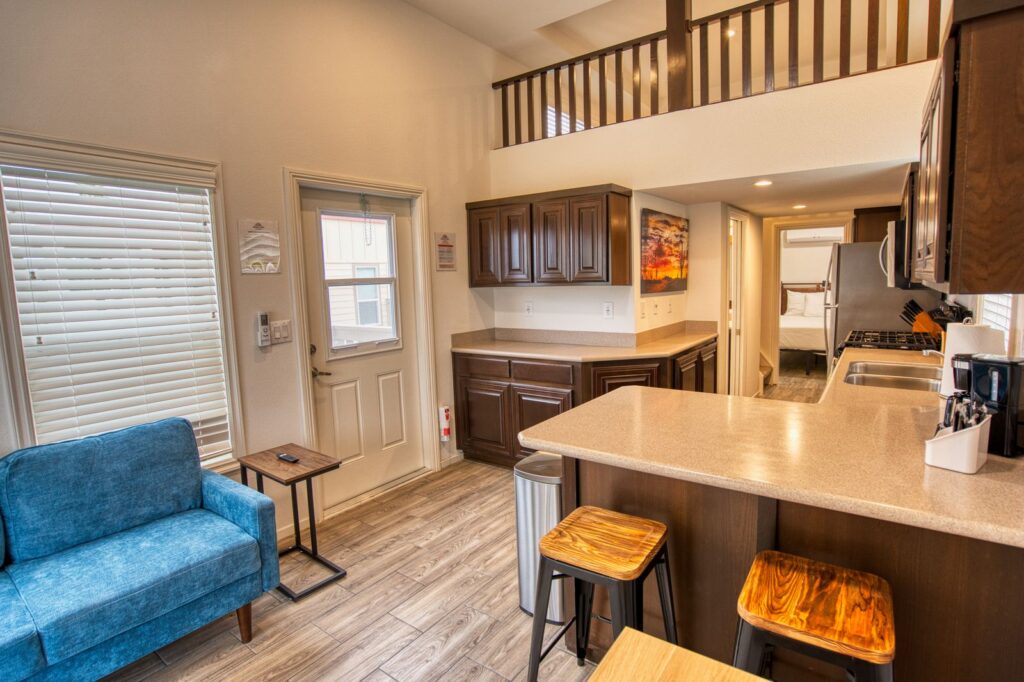 Open kitchen and living area with wood cabinets, breakfast bar, blue armchair, loft railing above, and visible bedroom through hallway. Natural light from windows and door on the left.