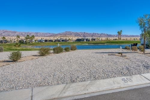 Gravel lot labeled 58 borders a small lake with palm trees, grass, and distant mountains under a clear blue sky.