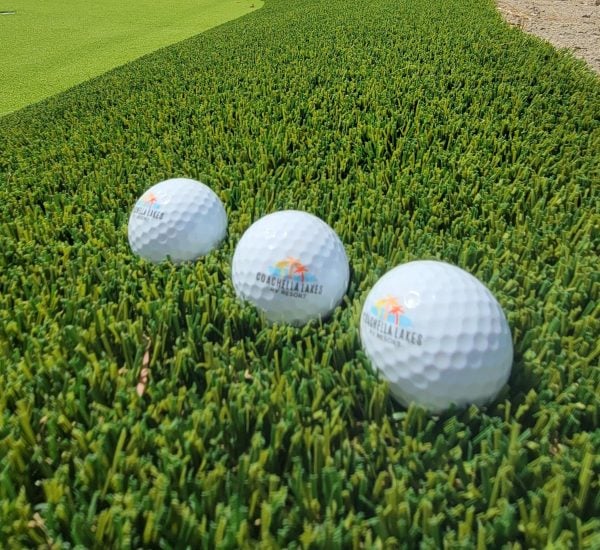 Three branded golf balls are lined up on artificial turf beside a putting green.