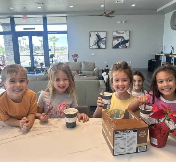 Five young children stand at a kitchen counter, smiling and holding cups of hot cocoa, with a hot cocoa mix box and a poinsettia plant on the counter.