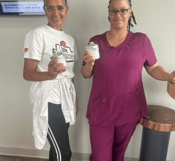 Two women stand indoors; one wears a white T-shirt and track pants, the other wears purple scrubs. Both are holding glass jars and smiling at the camera.