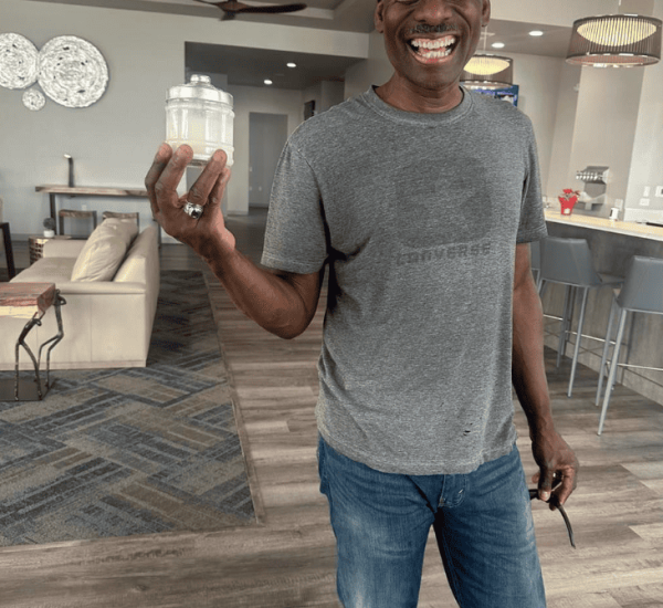A man in a gray t-shirt and jeans stands indoors, smiling and holding a jar with a white substance. The background shows a modern living area with tables, chairs, and decor.