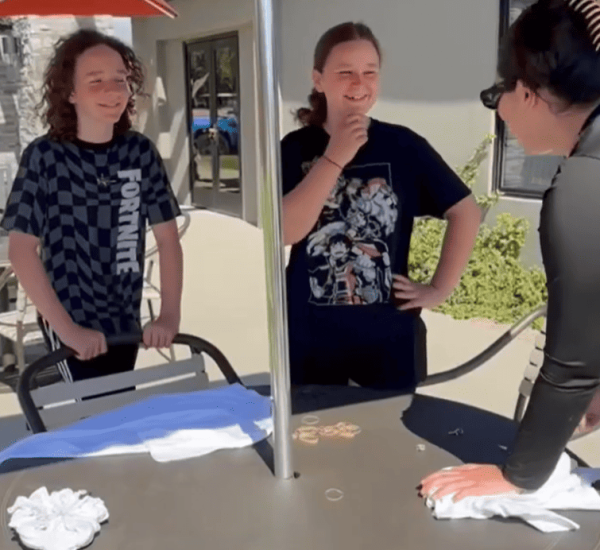 Two teenagers stand by a round outdoor table with white towels and coins while an adult speaks to them under a red umbrella.