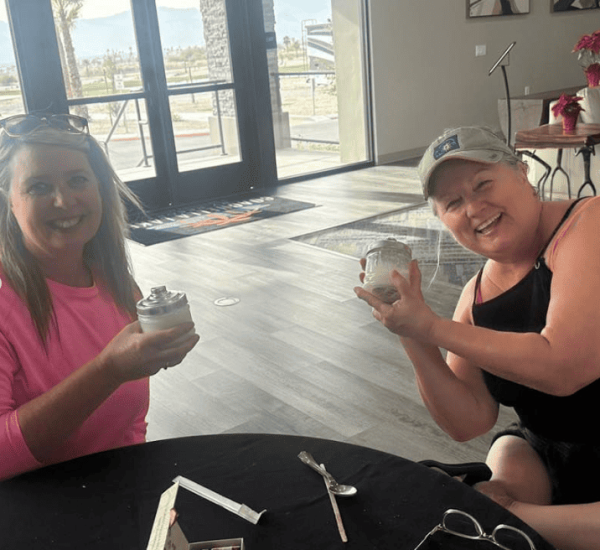Two women sitting at a table, each holding a dessert jar with whipped cream. There are utensils and a box on the table, and an open door with an outdoor view in the background.