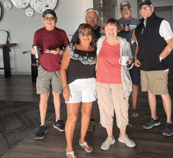 Six adults stand indoors, posing for a group photo. Most are dressed casually in shorts and T-shirts, and some hold coffee cups. The background features wall art and modern decor.